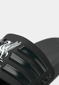 Fekete, műanyagból készült papucs, három fényes csíkkal és egy strukturált felsőrésszel, amelyen egy fehér szárnyas embléma található.