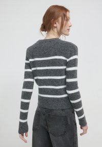 Femme aux cheveux roux de profil, portant un pull en laine gris avec des rayures horizontales blanches et un jean en denim noir, devant un fond uni.