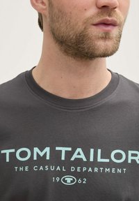 Pilkos spalvos marškinėliai su apvalia apykakle, ant kurių įspaustos šviesiai mėlynos spalvos raidės: "TOM TAILOR THE CASUAL DEPARTMENT 1962", aiškiai matomos priekyje.