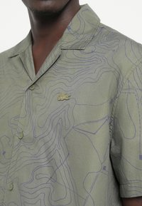 Short-sleeve shirt in olive green fabric featuring a blue contour line pattern and a small gold logo accent on the chest area.