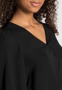 Blouse noire avec un décolleté en V, texture douce, détails de boutons et manches amples. Le tissu semble léger et tombe avec fluidité.