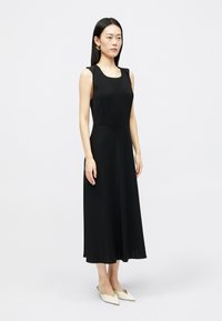 Γυναίκα που στέκεται φορώντας ένα αμάνικο μαύρο μίντι φόρεμα και κρέμα μοντέρνα παπούτσια με μυτερή μύτη, σε έναν απλό ανοιχτό φόντο.