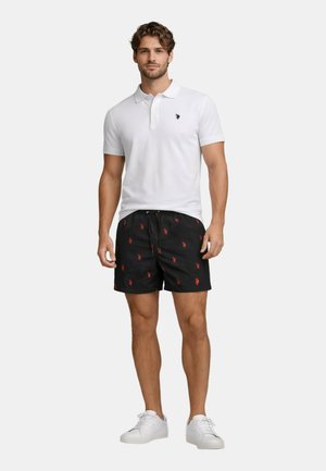 Mann trägt weißes kurzärmeliges Poloshirt, schwarze Shorts mit Kordelzug und roten Logos sowie weiße Turnschuhe, steht in entspannter Haltung.
