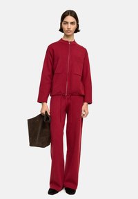 Czerwona kurtka zapinana na zamek z dwoma przednimi kieszeniami, w połączeniu z szerokimi spodniami i czarnymi butami wsuwanymi. Modelka trzyma ciemną zamszową torbę na ramię.