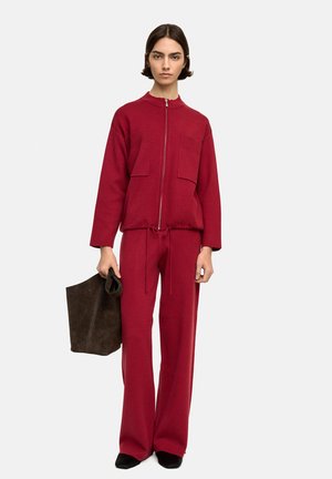 Czerwona kurtka zapinana na zamek z dwoma przednimi kieszeniami, w połączeniu z szerokimi spodniami i czarnymi butami wsuwanymi. Modelka trzyma ciemną zamszową torbę na ramię.