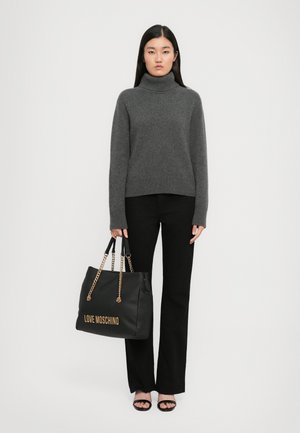 Woman wearing a gray turtleneck sweater, black pants, black heeled sandals, holding a large black Love Moschino handbag with gold chain straps.
