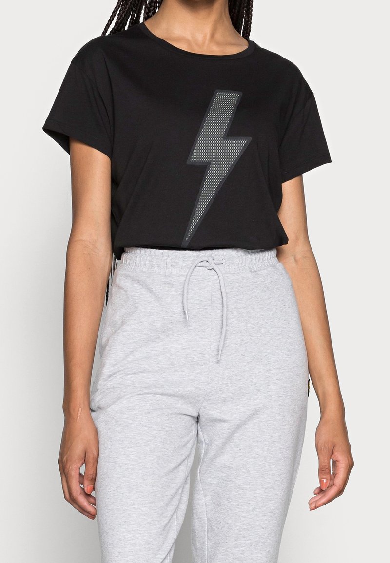 Personne portant un t-shirt noir à manches courtes avec un grand éclair gris, associé à un pantalon de survêtement gris clair à cordon.