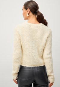 Un pull en tricot épais de couleur crème, avec des manches longues et une coupe légèrement cropped, associé à un pantalon en cuir noir ajusté.