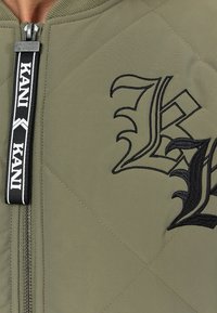 Giacca bomber trapuntata verde oliva con logo ricamato nero, chiusura con zip dotata di un tirante marchiato a contrasto in bianco e nero.