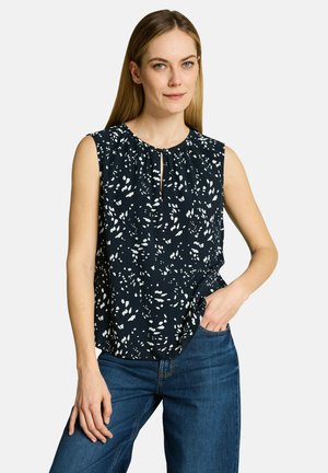 Frau mit glattem blondem Haar, die ein ärmelloses schwarzes Oberteil mit weißen Sprenkeln und blaue Jeans trägt, steht mit einer Hand in der Tasche.