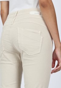 Beige high-waisted jeans with a textured fabric. Features two back pockets and a branded waistband label at the back.