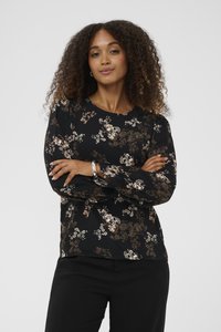Ženska s kodrasto frizuro, oblečena v črno cvetlično bluzo z dolgimi rokavi in črne hlače, stoji s prekrižanimi rokami pred enobarvno ozadjem.