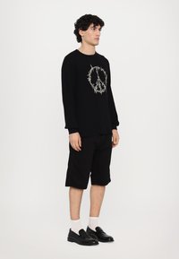Black long-sleeve sweatshirt with a peace symbol design in gray. Paired with black knee-length shorts and black slip-on shoes, accented with white socks.