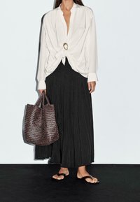 Blouse blanche avec un nœud à l'avant et un accent doré ; jupe longue plissée noire ; sac cabas tissé marron ; sandales plates.