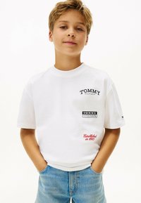 T-shirt in cotone bianco con collo rotondo, caratterizzata da loghi ricamati neri e rossi, e un dettagli di testo rosso "Fondato nel 1985."