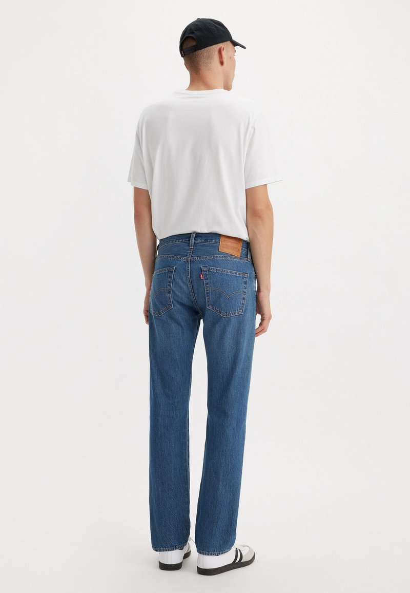 Blaue Denim-Jeans mit geradem Schnitt, gestickten hinteren Taschen und einem braunen Lederpatch am Bund. Getragen mit einem weißen T-Shirt.