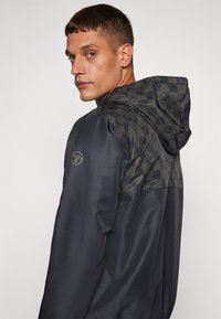 Chaqueta de un azul marino oscuro con capucha texturizada y a cuadros. Presenta un logo sutil en la manga. Tejido suave con una apariencia ligera.