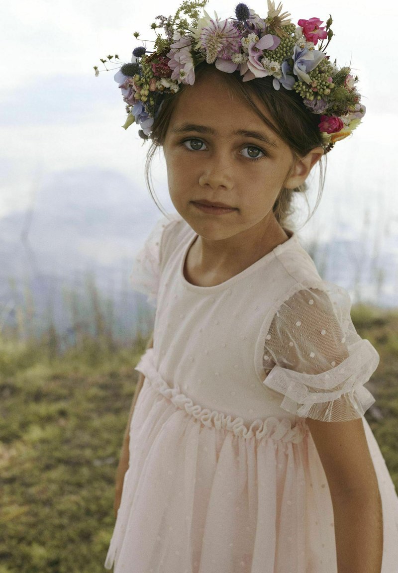 Robe rose clair avec des manches bouffantes courtes et transparentes, et une texture à pois. Couronne de fleurs avec un assortiment de fleurs. Détail de taille froncée douce.