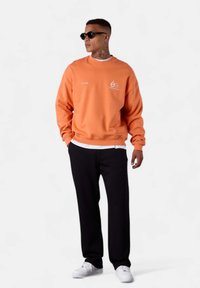 Sweatshirt orange avec un col rond, présentant un détail de logo blanc, associé à un pantalon noir et des baskets blanches, porté par un mannequin portant des lunettes de soleil.