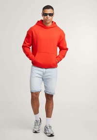 Felpa rossa con tasca a marsupio, abbinata a pantaloni corti in denim chiaro e strappati. Indossata con scarpe sportive grigie e blu e occhiali da sole neri.