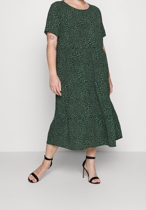 Nainen, jolla on vihreä mekko, jossa musta lehtikuvio ja lyhyet hihat, yhdistetty mustiin korkokenkäisiin nilkkaremmi-sandaaleihin.