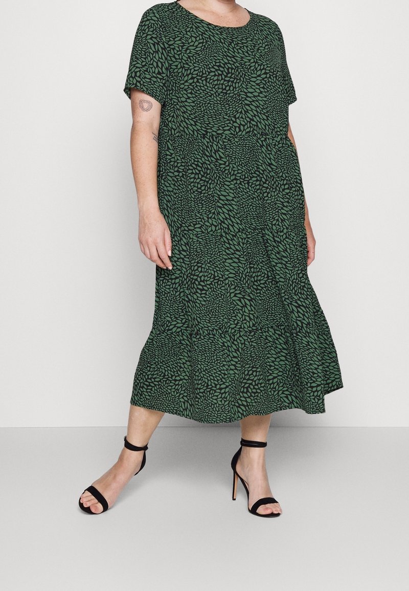 Femme portant une robe verte avec motif de feuilles noires et manches courtes, associée à des sandales noires à talons hauts avec bride à la cheville.