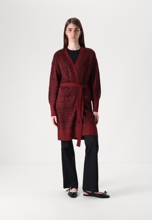 Cardigan rouge en tricot avec un motif complexe, devant ouvert, ceinture assortie et poignets côtelés, associé à un pantalon noir et des chaussures noires.