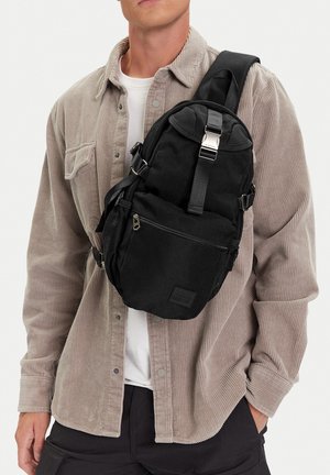 Muž v béžové košili z manšestru s černým crossbody batohem s několika kapsami a stříbrnou přezkou na bílém pozadí.