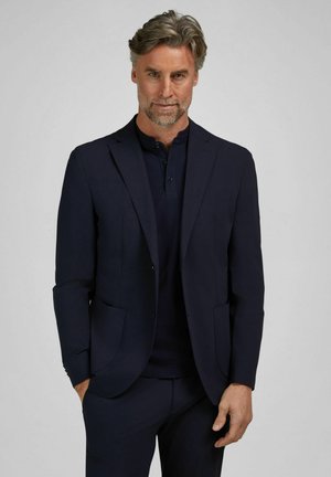 Man with gray hair and beard wears dark navy suit jacket and buttoned shirt, standing with one hand in pocket against plain background.