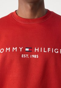 Czerwona bluza z okrągłym dekoltem. Posiada haftowany napis "TOMMY HILFIGER" oraz logo w kolorach białym, niebieskim i czerwonym. Tkanina jest gładka.
