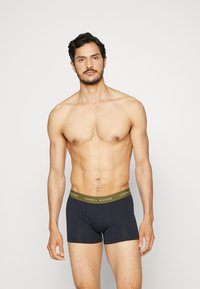 Črne bokserice z zelenim pasom, na katerem je v belem napisan "TOMMY HILFIGER". Gladek material, prilagojen dizajn, dolžina do sredine stegen.