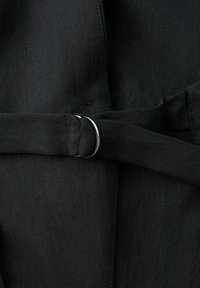 Ceinture en tissu noir avec boucle double en D argentée, attachée autour d’un vêtement noir assorti, montrant la texture et les détails de couture.
