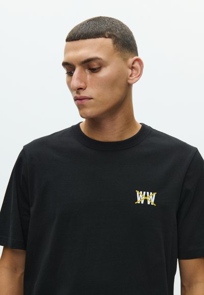 Μαύρο βαμβακερό μπλουζάκι με μικρό λευκό και κίτρινο σχέδιο στο αριστερό στήθος, με κοντά μανίκια και στρογγυλή λαιμόκοψη.