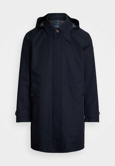 Casaquinho impermeável azul marinho com capuz, fecho de botão, dois bolsos frontais e detalhes em abas nas mangas. Acabamento exterior liso.