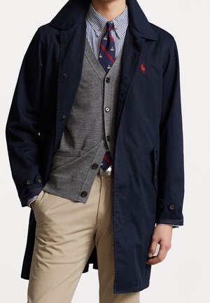 Mężczyzna ubrany w granatowy płaszcz na szarym, zapinanym swetrze, niebiesko-białą koszulę w paski, wzorzysty krawat i beżowe spodnie.