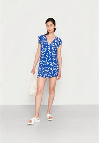 Vestido floral azul com mangas curtas, decote em V e bainha com babados. Com sandálias brancas e uma bolsa branca texturizada. Design simples.