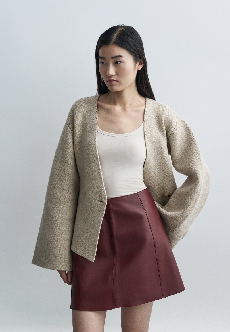 Smėlio spalvos vilnonis kardiganas su didelėmis rankovėmis, derantis prie aptemptos bordo odinės sijono. Lygios tekstūros su subtiliomis detalėmis.