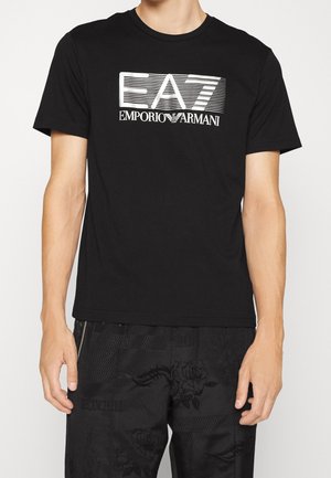 Mężczyzna w czarnej koszulce z białym logo „EA7 Emporio Armani” oraz czarnych spodniach z delikatnym kwiatowym wzorem.