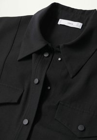 Chemise noire à col avec boutons-pression et deux poches poitrine sur fond blanc.
