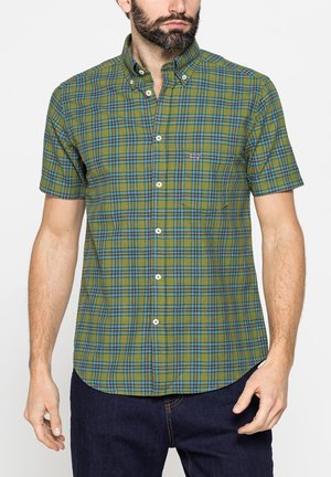 Uomo che indossa una camicia a maniche corte a quadri verdi e blu con colletto abbottonato e jeans blu scuro, in piedi davanti a uno sfondo semplice.