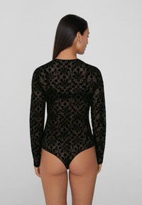 Czarny bodysuit z długimi rękawami, wykonany z przezroczystej tkaniny z teksturowanym wzorem damasku. Tył projektu podkreśla wysoko wykrojony nogawkę.