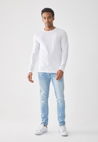 Langermet hvit skjorte med rund hals og en liten gul logo, sammen med lyseblå jeans og hvite sneakers. Glatt stoff, avslappet passform.