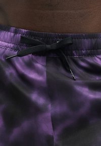 Κοντινό πλάνο από μοβ και μαύρα σορτς tie-dye με μαύρο κορδόνι δεμένο σε φιόγκο στη ελαστική μέση πάνω σε σκούρο δέρμα.