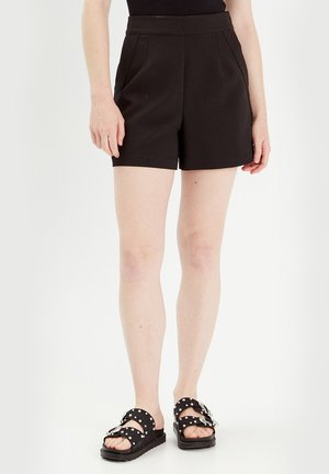 Shorts noirs sur mesure avec des plis, une taille haute, et un tissu lisse. Associés à des sandales noires à motif à pois et à boucles.