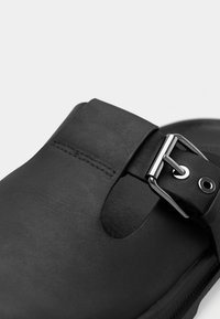 Μαύρο δερμάτινο σανδάλι με λουράκι με αγκράφα, κοψίματα στο πάνω μέρος και λείο υλικό στην εσωτερική σόλα. Η σόλα έχει ελαφριά καμπυλότητα.