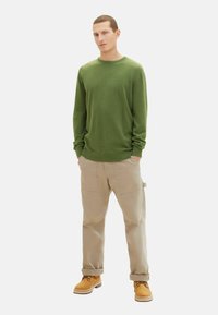 Grön stickad tröja,beige cargobyxor och bruna stövlar. Tröjan har en ribbad rund halsringning och långa ärmar. Cargobyxorna har sidofickor.