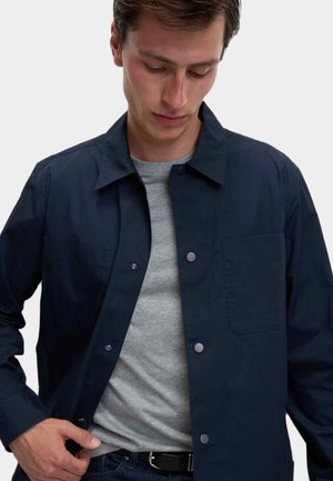 Uomo con giacca blu navy con bottoni sopra una t-shirt grigia, che allaccia la giacca con una mano, guardando verso il basso.
