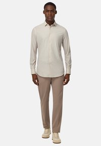 Camisa beige de manga larga con botones y cuello, combinada con pantalones beige claros y zapatillas color crema, de pie frente a un fondo liso.