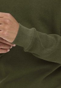Maglione a maglia verde khaki, tessuto testurizzato, polsini a costine, primo piano dell'area del polso, motivo di intreccio morbido, senza loghi o decorazioni visibili.