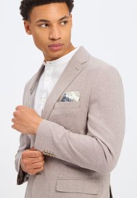 Lichtbeige blazer van gestructureerde stof, met een enkele borstzak voorzien van een patroonaccesoire en subtiele knoopaccenten.